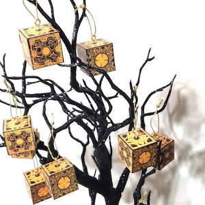 May include: A black tree branch with seven gold ornaments shaped like a cube with a complex pattern. The ornaments are hanging from the branches. The tree is in a black pot.