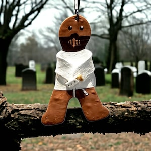 May include: A brown gingerbread man ornament with a white straitjacket, hanging from a branch in a cemetery.
