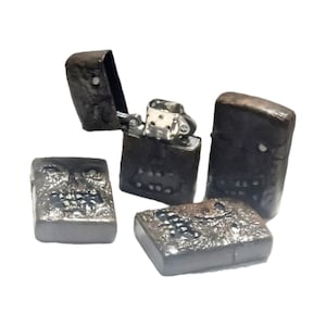 May include: Four rectangular metal lighters with a dark, textured, weathered finish. One lighter is open, revealing the silver-colored internal mechanism. Each lighter features a sculpted, skull-like face design with visible eye sockets and teeth.