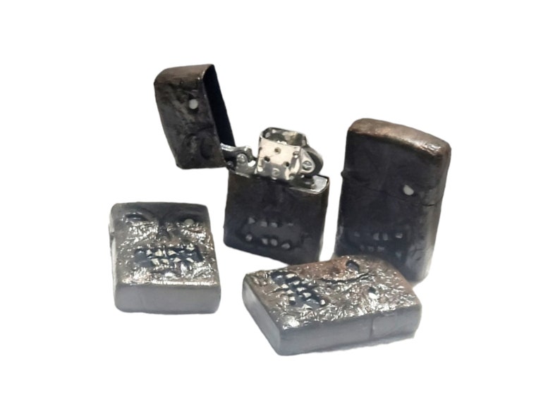 May include: Four rectangular metal lighters with a dark, textured, weathered finish. One lighter is open, revealing the silver-colored internal mechanism. Each lighter features a sculpted, skull-like face design with visible eye sockets and teeth.