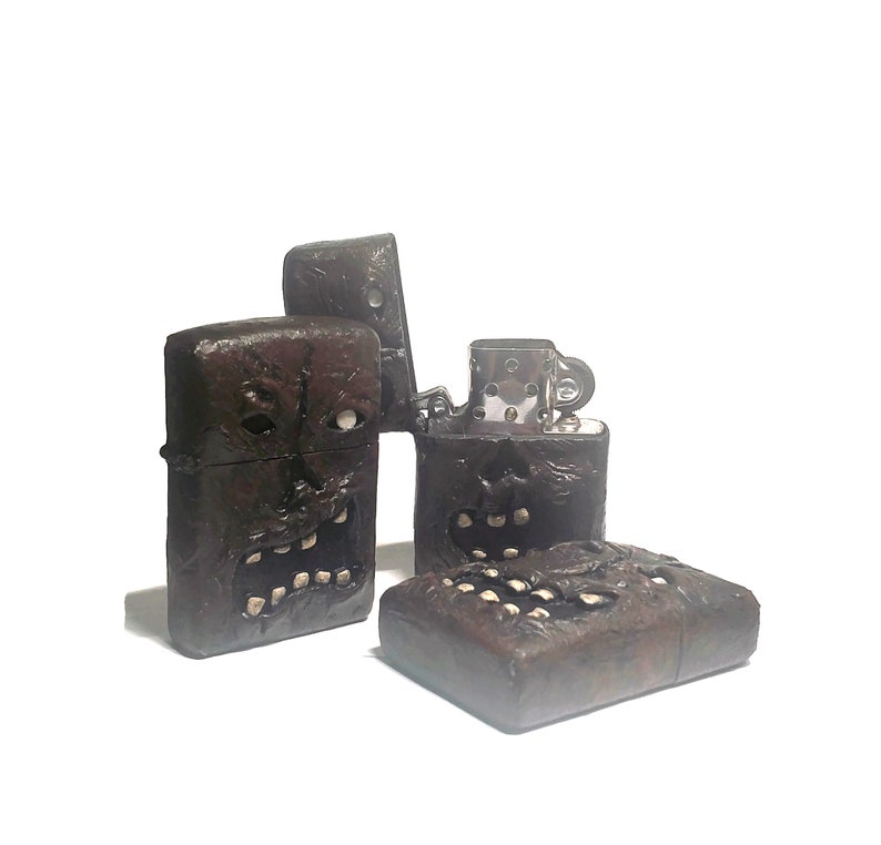 May include: Two dark brown metal lighters with a textured, sculpted surface resembling a monstrous face with visible teeth and eyes. One lighter is standing upright with its lid open, revealing the silver-colored internal mechanism, while the other lies flat.