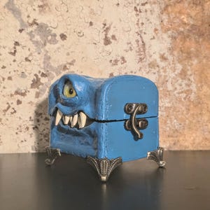 May include: A blue monster-themed treasure chest with a single eye, sharp teeth, and decorative feet. The chest has a hinged lid and a metal clasp. The background is a textured, mottled wall.