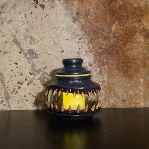 May include: A navy blue, handcrafted lantern with a unique design. The lantern features a glass enclosure with a row of sharp, tooth-like protrusions around the middle. A yellow candle is lit inside. The lantern sits on a black surface.