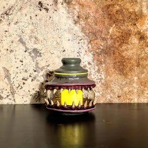May include: A decorative jar with a unique design. The jar has a dark green lid and a clear glass body. The rim is outlined in purple and features a row of white teeth. The interior glows with a yellow light, creating a spooky effect.