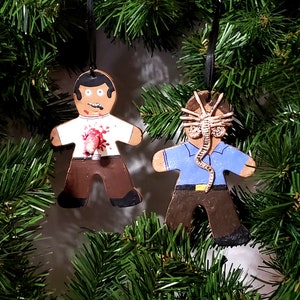 May include: Two gingerbread man ornaments with black ribbon hangers. One has a zombie design with a bloody chest. The other has an alien facehugger design. Both are brown, white, and blue, and are set against a backdrop of green pine needles.