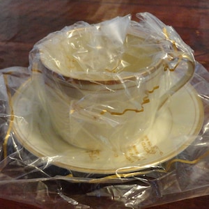 May include: A cream-colored teacup and saucer set with gold trim, wrapped in clear plastic. The cup has a handle and sits on a matching saucer. The set is on a wooden surface.
