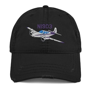 Könnte beinhalten: Eine schwarze, distressed Baseballkappe mit einer Stickerei eines zweimotorigen Flugzeugs in Weiß, Blau und Lila. Über dem Flugzeug ist der Text N1903 in lilafarbenem Garn gestickt. Die Kappe hat einen gebogenen Schirm und eine Sechs-Panel-Konstruktion.