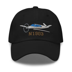 Könnte beinhalten: Eine schwarze Baseballkappe mit einer Stickerei eines weißen und grauen Flugzeugs mit blauen Fenstern. Unter dem Flugzeug ist der Text N1903 in braunem Garn gestickt. Die Kappe hat einen gebogenen Schirm und eine strukturierte Krone mit Ösen.