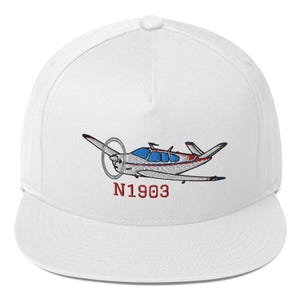 May include: A white baseball cap featuring an embroidered graphic of a small propeller airplane in white, blue, and red. Below the airplane, the text N1903 is embroidered in red thread. The cap has a structured crown and a flat brim.