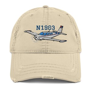 Könnte beinhalten: Eine beige, distressed Baseballkappe mit einer Stickerei eines Leichtflugzeugs in Weiß, Blau und Rot. Über dem Flugzeug ist der Text N1903 in Blau gestickt. Die Kappe hat einen gebogenen Schirm und eine abgenutzte, ausgefranste Optik.