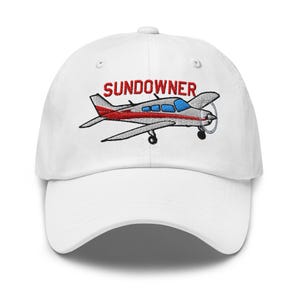 Könnte beinhalten: Weiße Baseballkappe mit einem gestickten Flugzeugdesign und dem Wort "SUNDOWNER" in Rot. Das Flugzeug ist silberfarben mit roten Akzenten und blauen Fenstern. Eine klassische, verstellbare Kappe für Luftfahrtbegeisterte.