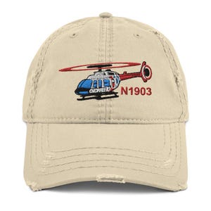 May include: A beige distressed baseball cap featuring an embroidered graphic of a blue and red helicopter with the text N1903 stitched in red thread to the right of the aircraft. The cap has a curved brim and a six-panel construction.