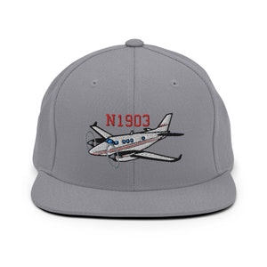 Könnte beinhalten: Graue Cap mit flachem Schirm und gesticktem Flugzeugdesign. Das Design enthält den Text "N1903" in Rot über dem Flugzeug. Das Flugzeug ist weiß mit roten und blauen Akzenten.