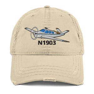 Könnte beinhalten: Eine beige Baseballkappe im Used-Look mit einer Stickerei eines weißen und blauen Leichtflugzeugs im Flug. Unter dem Flugzeug ist der Text N1903 in schwarzem Garn gestickt. Die Kappe hat einen gebogenen Schirm und eine Konstruktion aus sechs Teilen.