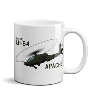 May include: A white ceramic mug featuring a graphic illustration of a dark olive green Boeing AH-64 Apache attack helicopter in flight. The text BOEING AH-64 is printed in black above the helicopter, and the word APACHE is printed below it.