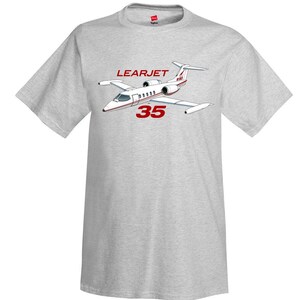 May include: A light gray short-sleeve t-shirt featuring a graphic of a white Learjet 35 aircraft with red accents. The text "LEARJET" appears in red above the plane, and the number "35" is printed in large red font directly below the aircraft.