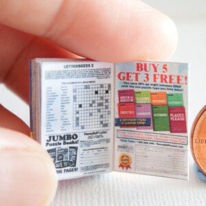 May include: A miniature open book with crossword puzzles and advertisements, held near a penny for scale. The book's pages display text including "JUMBO Puzzle Books!" and "BUY 5 GET 3 FREE!" The book is blue and white.