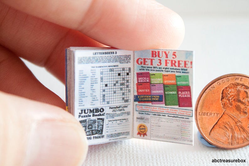May include: A miniature open book with crossword puzzles and advertisements, held near a penny for scale. The book's pages display text including "JUMBO Puzzle Books!" and "BUY 5 GET 3 FREE!" The book is blue and white.
