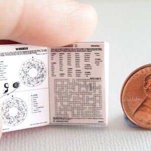 May include: A miniature open book with printed pages, featuring crossword puzzles and diagrams, held near a penny. The book's pages display text and illustrations. The penny is a copper color and shows the year 2015.