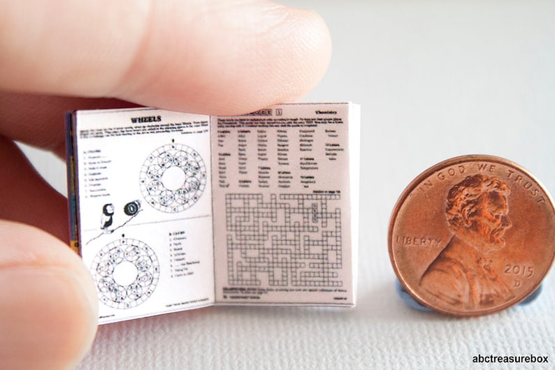 May include: A miniature open book with printed pages, featuring crossword puzzles and diagrams, held near a penny. The book's pages display text and illustrations. The penny is a copper color and shows the year 2015.