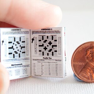 May include: A miniature open book of crossword puzzles with black and white grids and text, held near a penny. The book's pages display crossword grids and the words "Codeword" and "Family Ties". The penny is dated 2015.