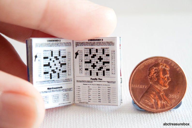 May include: A miniature open book of crossword puzzles with black and white grids and text, held near a penny. The book's pages display crossword grids and the words "Codeword" and "Family Ties". The penny is dated 2015.