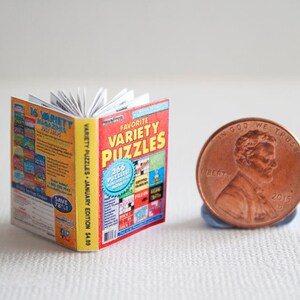 May include: A miniature book titled "Favorite Variety Puzzles" with a red and yellow cover, next to a US penny. The book is open, revealing white pages. The cover text includes "266 puzzles" and "January Edition".