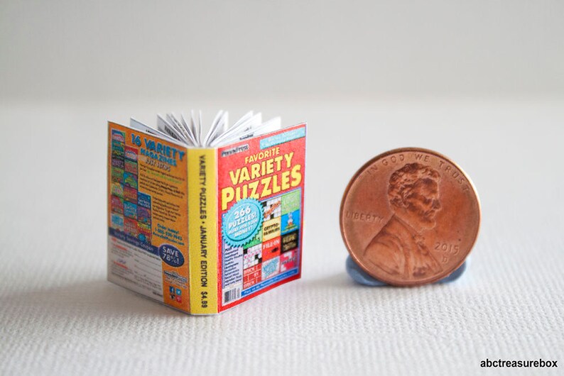 May include: A miniature book titled "Favorite Variety Puzzles" with a red and yellow cover, next to a US penny. The book is open, revealing white pages. The cover text includes "266 puzzles" and "January Edition".