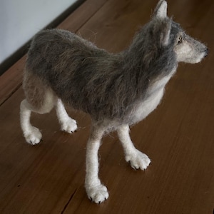 May include: A needle felted gray wolf with white legs and paws. The wolf is standing on a wooden surface.