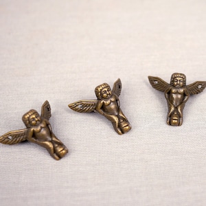 May include: Four antique gold-toned metal corner brackets shaped like cherubs with wings. Each bracket has a detailed design and is perfect for adding a decorative touch to furniture.