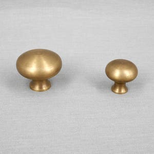 Pode incluir: Dois botões de armário de latão, um maior que o outro, com a parte superior arredondada e uma haste curta. Os botões estão sobre um fundo cinza claro.
