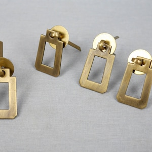 May include: Four gold-toned cabinet knobs with a rectangular shape and a square opening in the centre. The knobs have a simple, modern design.