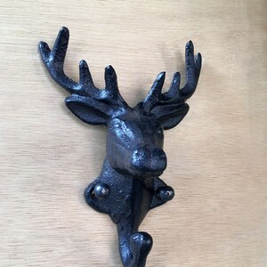 Può includere: Ganci da parete a forma di testa di cervo in ghisa nera con un unico gancio che si estende dalla parte inferiore. La testa di cervo è dettagliata con corna e un viso realistico.