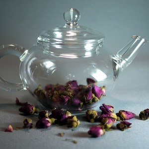 Puede incluir: Una tetera de vidrio transparente llena de capullos de rosa secos. La tetera tiene un cuerpo redondo y un asa curva. Los capullos de rosa son de color morado oscuro.