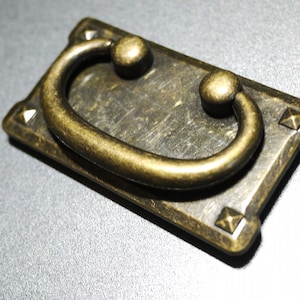 Puede incluir: Un tirador de cajón rectangular de color dorado antiguo con una manija redondeada en forma de D. El tirador tiene acentos decorativos en las esquinas.