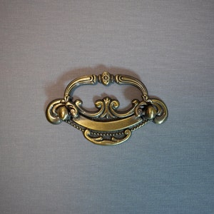 May include: An ornate drawer pull in a gold tone with a decorative scroll design.