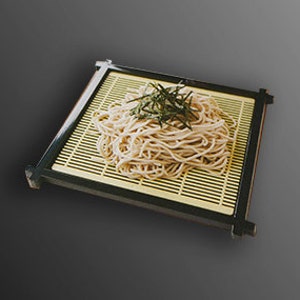 Könnte beinhalten: Nahaufnahme von einer Portion weißer Nudeln auf einer schwarzen und gelben Bambusmatte. Die Nudeln sind mit einer kleinen Menge grünem Seetang garniert.