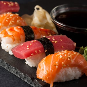 May include: A variety of sushi rolls on a black slate plate. The rolls include salmon, tuna, shrimp, and avocado. There is also a small dish of soy sauce and a small dish of wasabi.