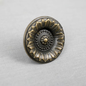 Könnte beinhalten: Ein runder, verzierter Knopf in Messingfarbe mit einem erhabenen, kreisförmigen Design in der Mitte.