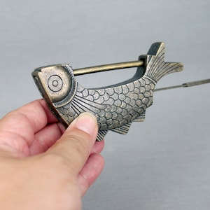 Op de afbeelding: Een messingkleurig slot in de vorm van een vis met een sleutelgat en een gedetailleerd ontwerp van vissen schubben. Het slot is open en de sleutel is aan het slot bevestigd.