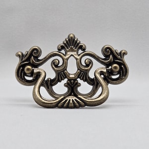 May include: A decorative, ornate, gold-toned metal drawer pull with a floral design.