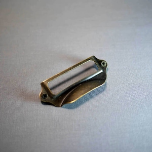 以下が含まれることがあります： 長方形のラベルホルダーが付いた真鍮製の引き出しの取っ手。取っ手は湾曲した形状で、2本のネジが見える。