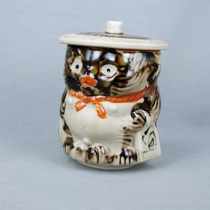 May include: A ceramic jar with a lid shaped like a hat. The jar is decorated with a brown and white cat wearing a red and orange bow tie.