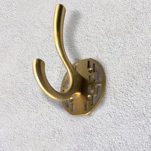 Puede incluir: Un gancho de metal dorado con un diseño geométrico decorativo. El gancho tiene una forma curva y está unido a una base circular.