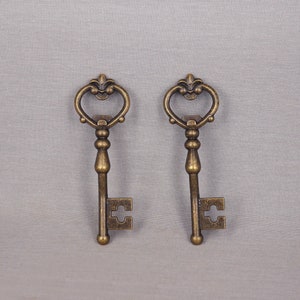 Peut inclure: Deux clés de style antique avec des sommets ornés en forme de cœur et des détails complexes. Les clés sont en métal de couleur bronze et sont présentées sur un fond gris neutre. Les clés sont décoratives.