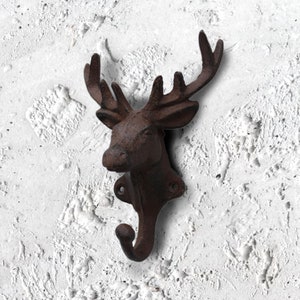 Puede incluir: Un gancho de pared de cabeza de ciervo de hierro fundido marrón con un solo gancho que se extiende desde la parte inferior. El gancho está montado en una pared blanca texturizada.