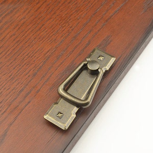 May include: A bronze-colored cabinet pull with a rectangular base and a ring-shaped handle. The pull has a decorative design with small stars on the corners.