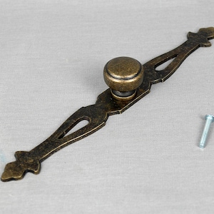 May include: A bronze drawer pull with an ornate design and a round knob. It comes with a silver screw.