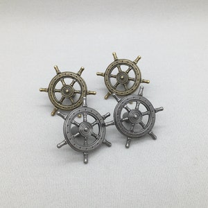 Op de afbeelding: Vier decoratieve scheepswielknoppen in antiek messing en zilver afwerkingen. Elk wiel heeft acht spaken en een centrale naaf. De knoppen zijn gerangschikt op een witte ondergrond, ideaal voor nautische decoratie.