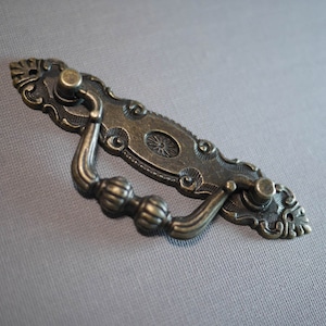 May include: Antique-style bronze drawer pull with ornate details and a decorative knob on each end.